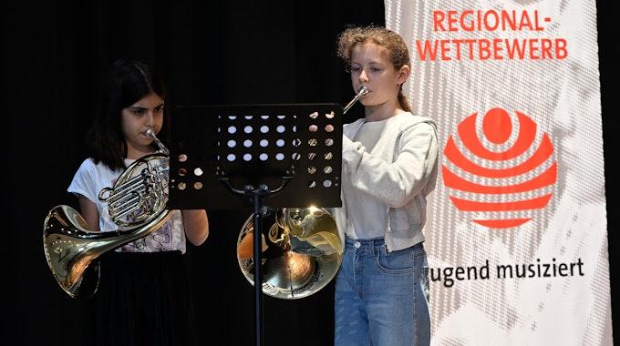 Die beiden stehen auf einer Bühne mit ihren Instrumenten, links im Bild ein Banner.