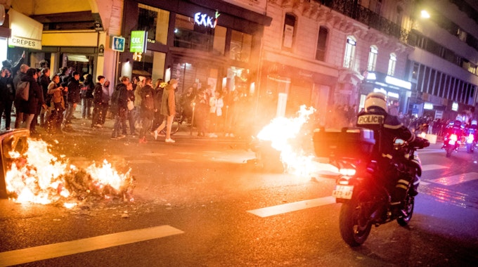 Ein Polizist fährt auf einem Motorrad an brennenden Mülltonnen in der französischen Hauptstadt Paris vorbei. In der Nacht zu Dienstag sind aufgrund der verabschiedeten Rentenreform Hunderte Menschen auf die Straßen gegangen.