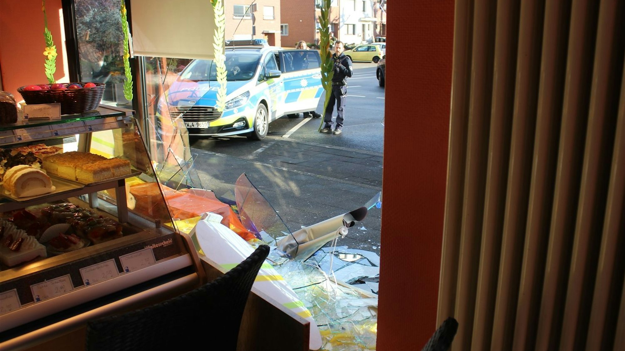 Das Foto zeigt eine zerstörte Glasscheibe und einen Polizeiwagen vor der Bäckerei.