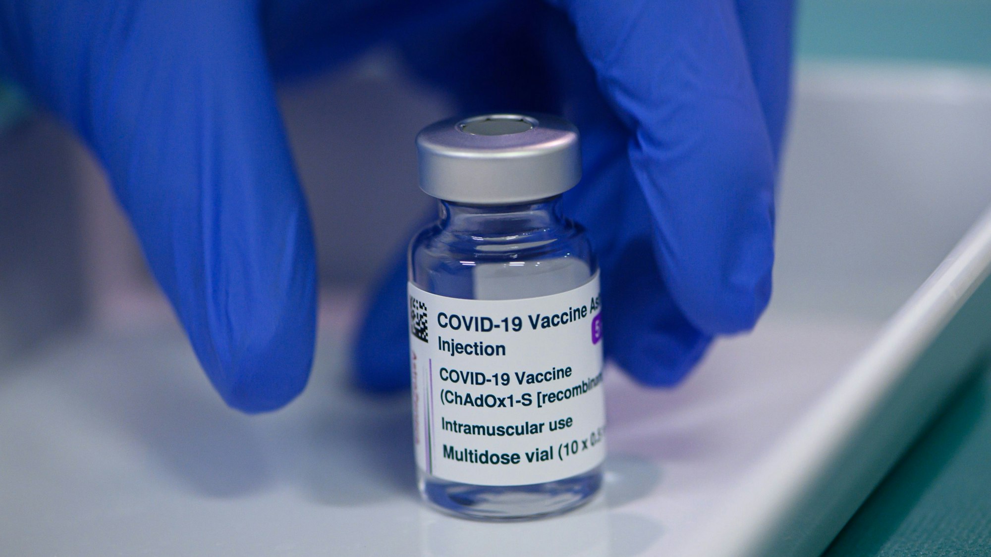 Eine Ampulle des Impfstoffs Vaxzevria vonAstrazeneca, die von einer Hand mit einem blauen Handschuh genommen wird.