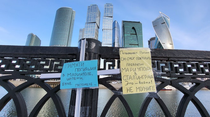Zettel in den ukrainischen Nationalfarben (blau und gelb) hängen an der Brücke des Flusses Moskwa in Moskau. Im Hintergrund sind Gebäude des Hochhausviertels Moskwa City zu sehen.