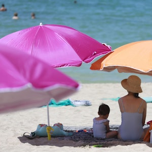 Eine Frau sitzt neben ihrem Kind unter einem Sonnenschirm an einem warmen Sommertag am Strand, hier im August 2022 auf Mallorca.