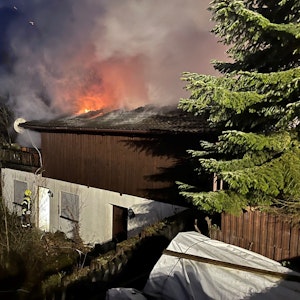 Auf dem Dach des Hauses sind Flammen zu sehen, vor der Tür steht eine Einsatzkraft der Feuerwehr.
