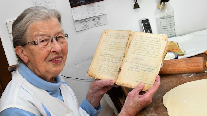 Oma Agnes sitzt an ihrem Küchentisch und zeigt ihr Rezeptbuch. Auf der Tischplatte liegen ausgerollter Teig und ein Nudelholz.