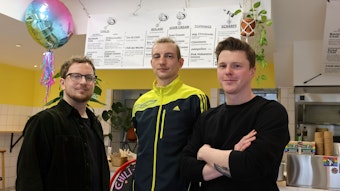 Drei junge Männer stehen in einem kleinen Laden vor einer Speisekarte an der Wand, daneben ist ein bunter Luftballon.