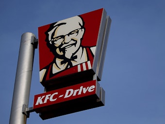 Das Symbolfoto aus dem Jahr 2018 zeigt einen Metall-Mast, an dem zwei große Schilder des US-Fast-Food-Konzerns Kentucky Fried Chicken befestigt sind. Im Hintergrund ist blauer Himmel zu sehen.