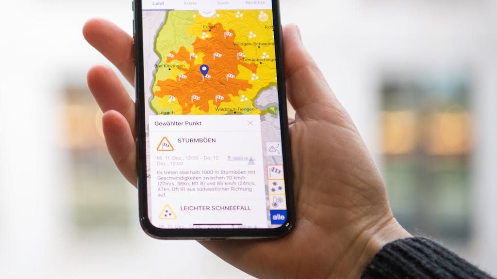 Das Symbolfoto aus dem Jahr 2019 zeigt eine Hand, die ein Smartphone hält. Auf dem Bildschirm sieht man eine Wetterkarte in gelb-orange, darunter steht Text.