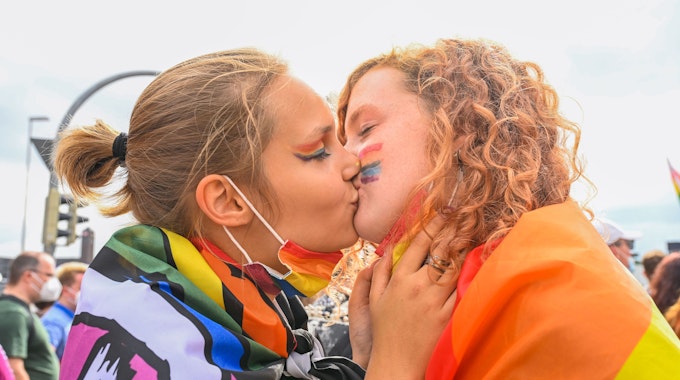 Das Symbolfoto aus dem Jahr 2021 zeigt zwei junge Frauen, die sich küssen. Beide sind in Regenbogen-Flaggen gehüllt.