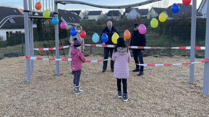 EInige Personen und Kinder stehen um ein mit Luftballons und Absperrband dekoriertes Spielplatzgerät herum.