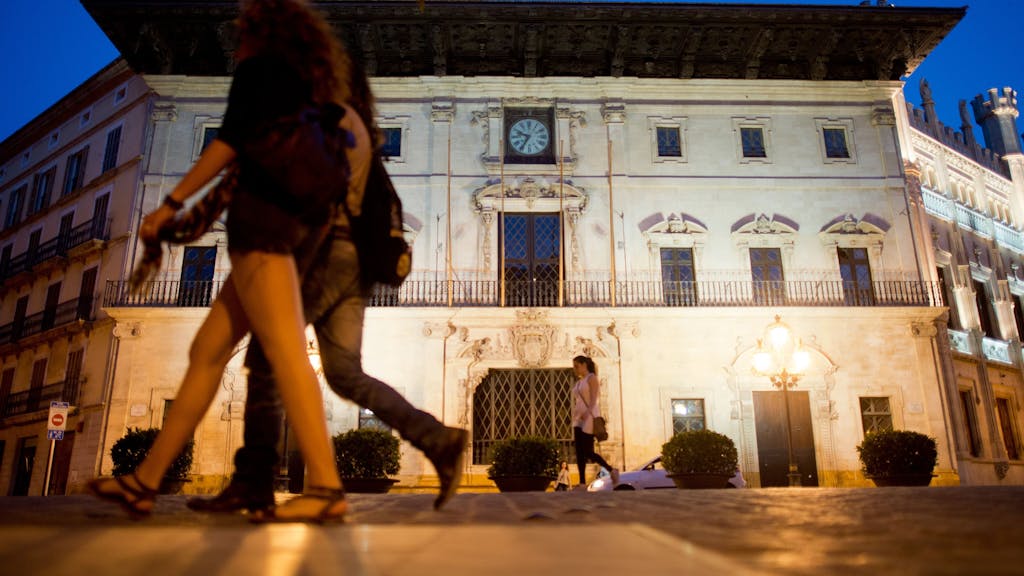 Das Bild zeigt das Rathaus in Palma de Mallorca bei Nacht. Zwei Passanten laufen im Vordergrund vorbei.