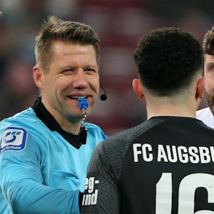 Schiedsrichter Patrick Ittrich lächelt im Gespräch mit Augsburgs Ruben Vargas. Neben ihm gestikuliert Kapitän Jonas Hector vom 1. FC Köln.