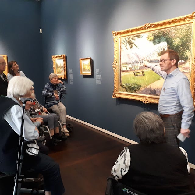 Ein Mann beschreibt ein Kunstwerk in einem Museum. Seniorinnen und Senioren schauen ihm zu.