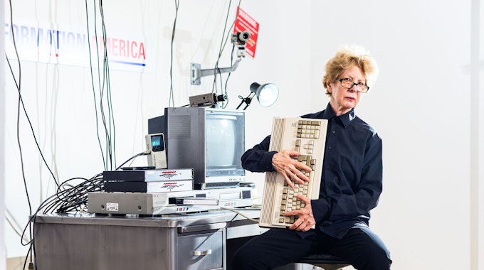 Julia Scher sitzt in ihrer Installation Information America und hält eine alte Computertastatur in den Händen.
