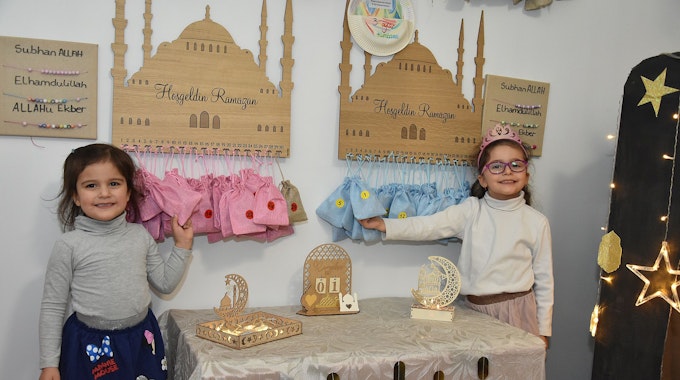 Zwei kleine Mädchen stehen vor Ramadan-Dekorationen. In dreißig Säckcken, die unter einem hölzernen Moschee-Schild aufgehangen sind, erwarten sie Geschenke.