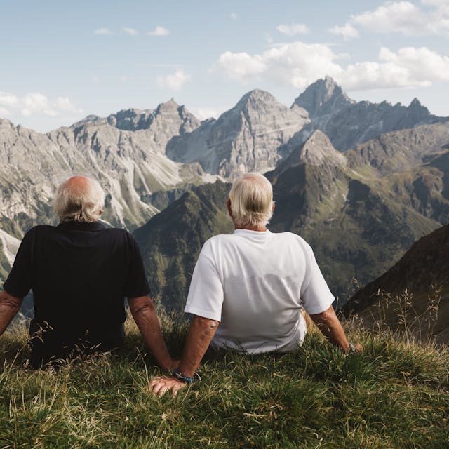 Zwei ältere Menschen sitzen an einem Hang und schauen auf eine Berglandschaft hinaus.