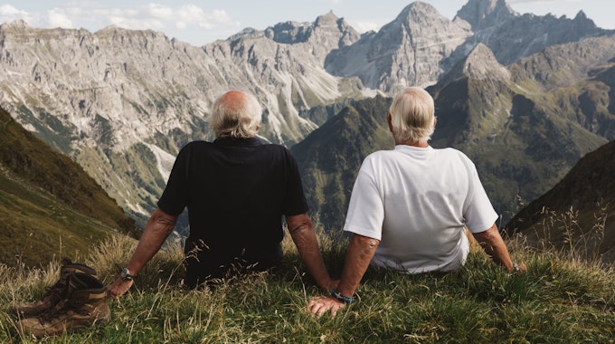 Zwei ältere Menschen sitzen an einem Hang und schauen auf eine Berglandschaft hinaus.