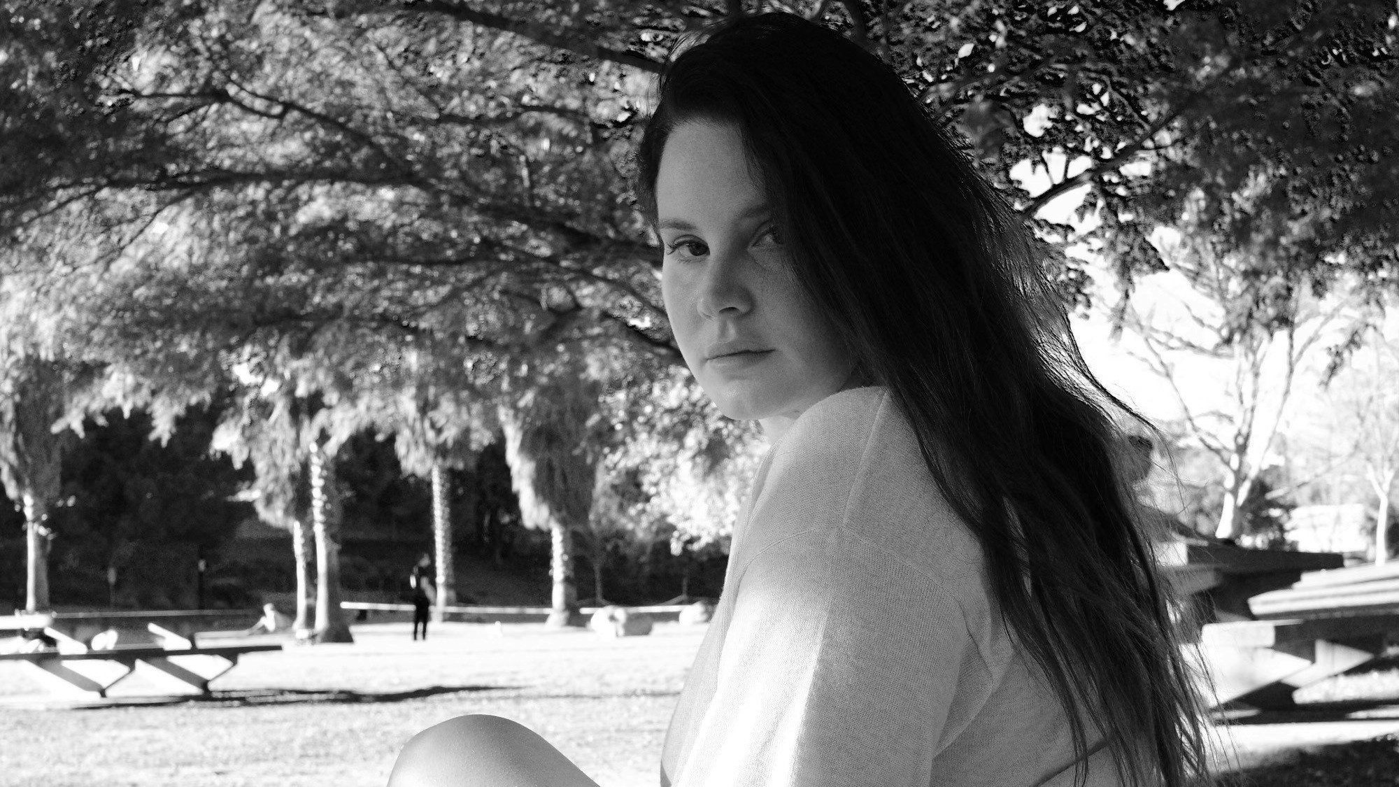 Lana Del Rey sitzt in einem Park unter einem Baum und schaut in die Kamera. Das Bild ist schwarz-weiß.
