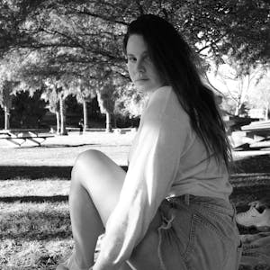 Lana Del Rey sitzt in einem Park unter einem Baum und schaut in die Kamera. Das Bild ist schwarz-weiß.