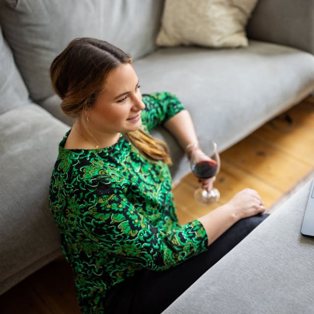 Eine Frau sitzt mit einem Glas Rotwein vor ihrem Laptop.&nbsp;