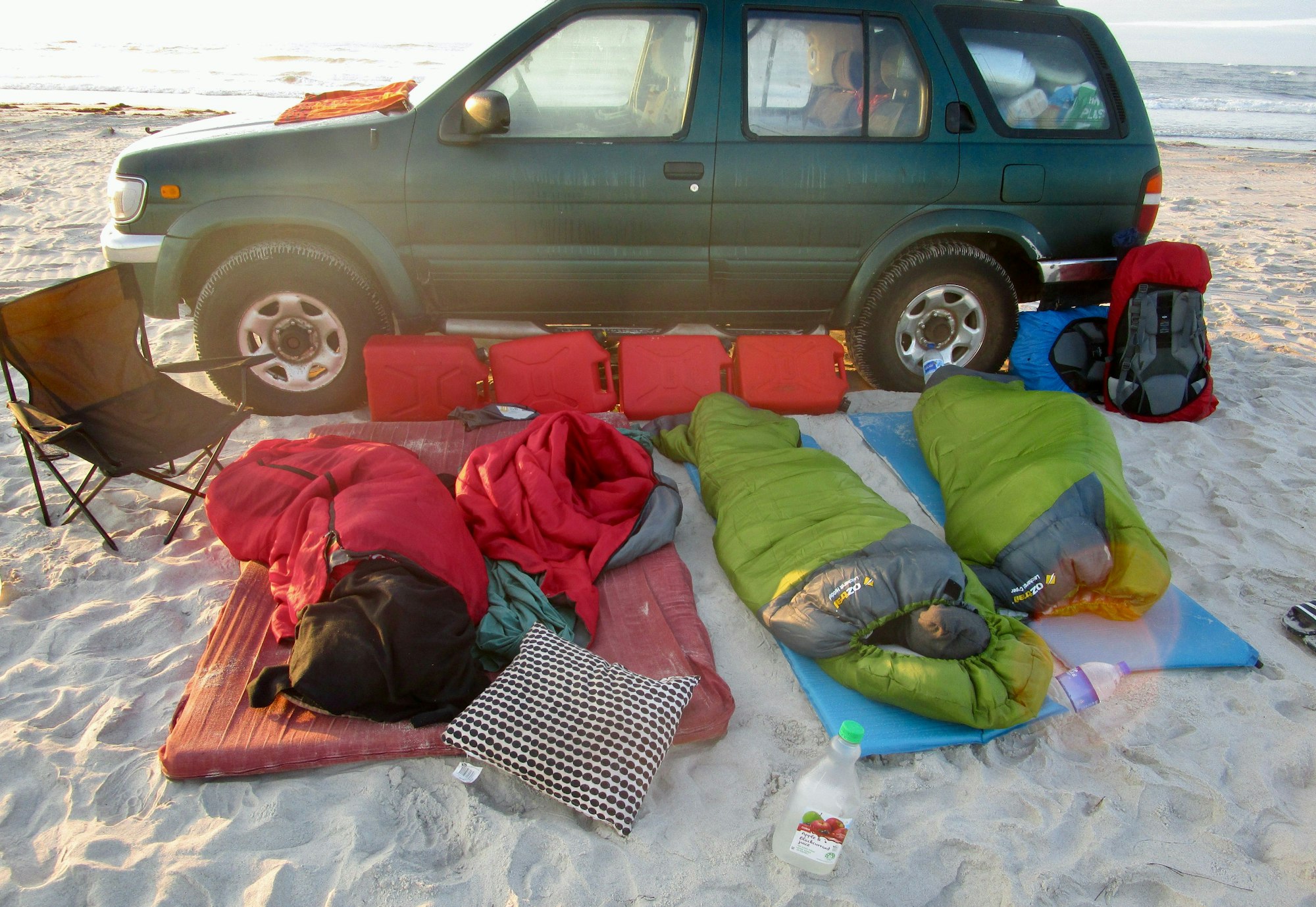 Ein Auto steht am Strand, davor schlafen zwei junge Menschen in einem Schlafsack.