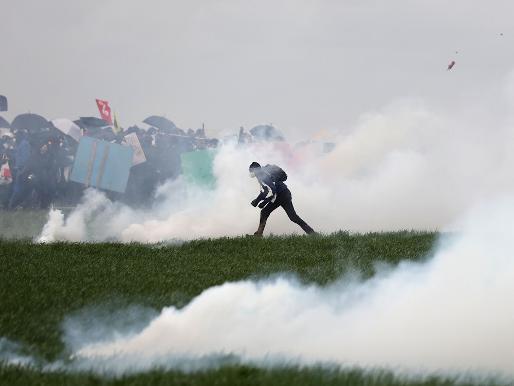 Ein Demonstrant steht inmitten von Tränengas auf dem Feld. Im Hintergrund sind weitere Demonstranten zu erkennen.