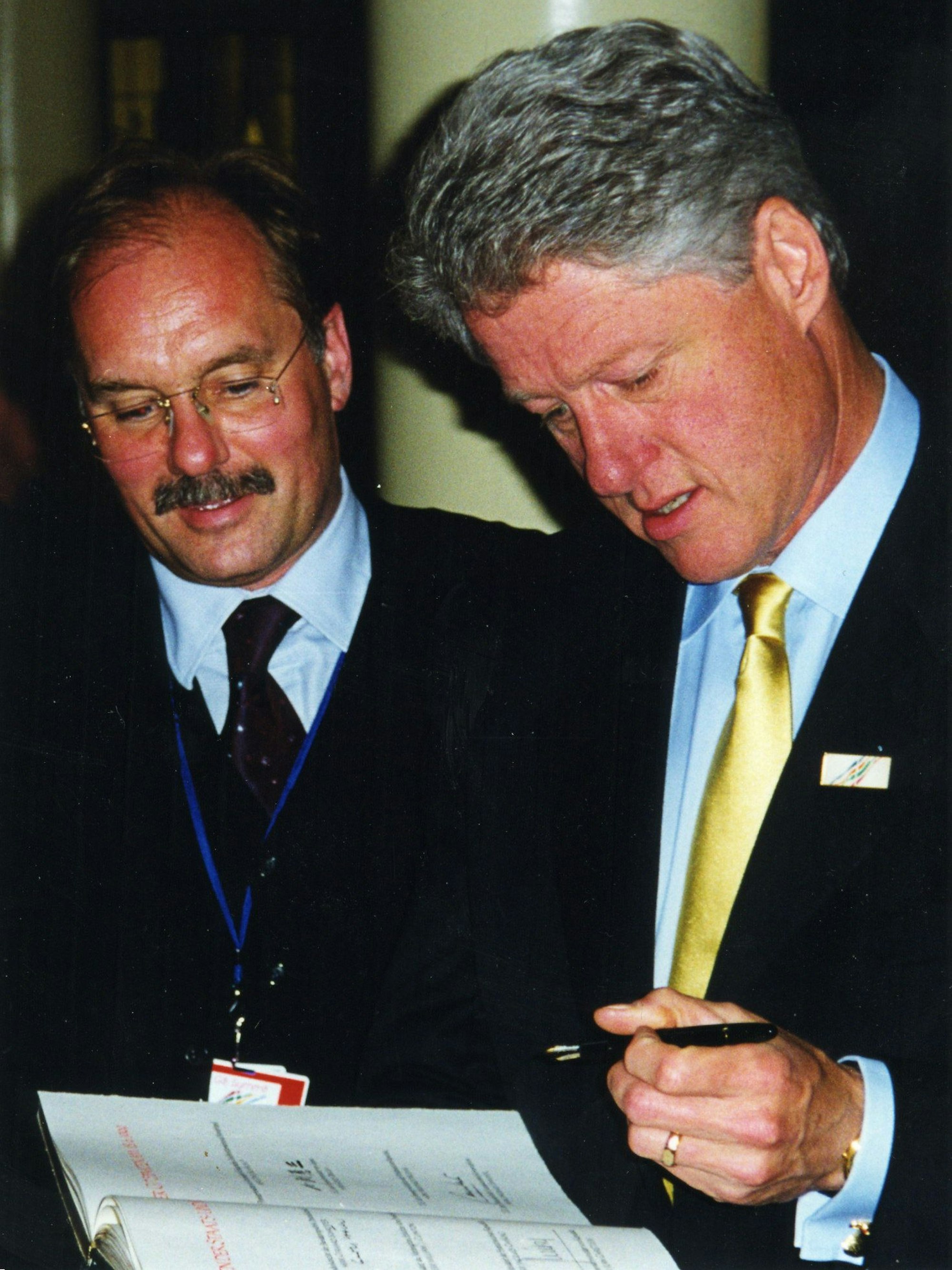 Bill Clinton hält ein Buch und einen Füller in den Händen, neben ihm steht Bernhard Conin.