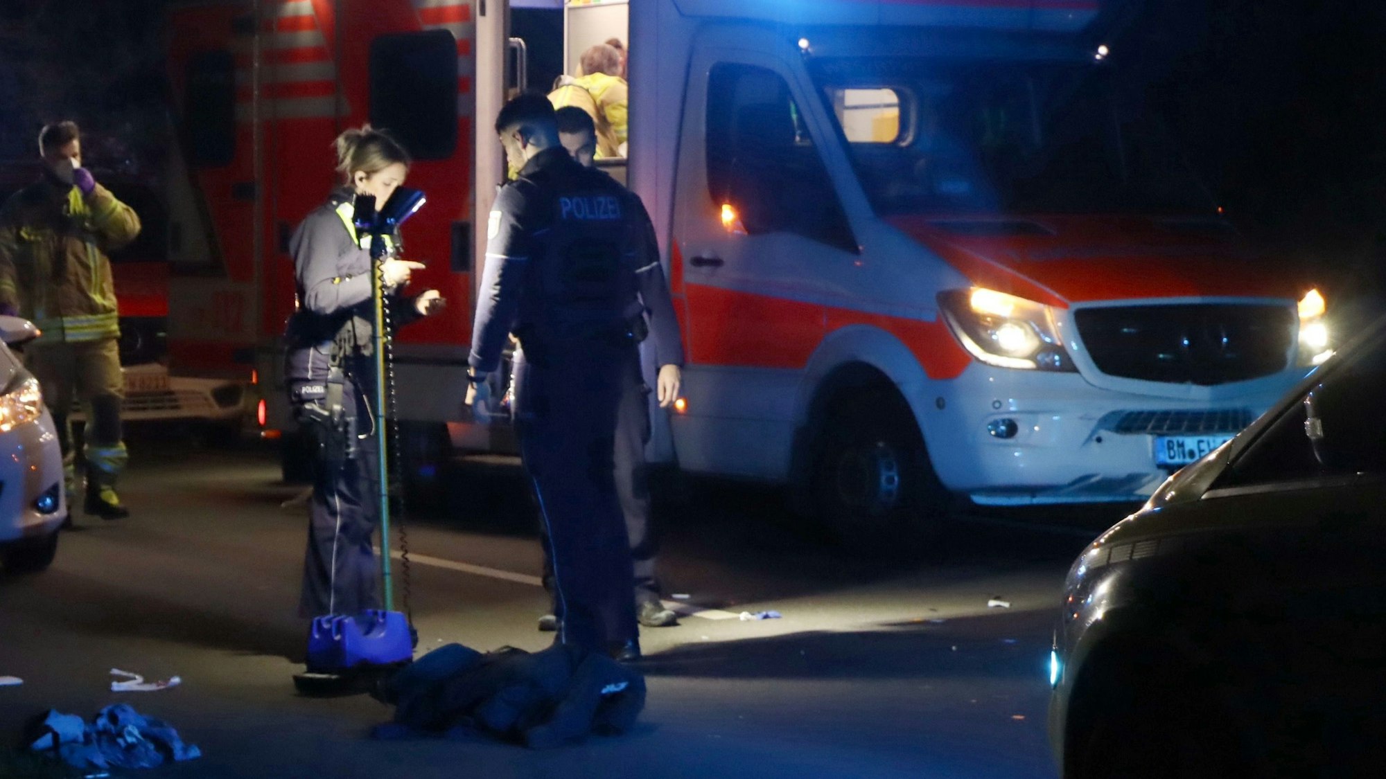 Auf dem Foto ist die hell erleuchtete Unfallstelle auf der Rodenkirchener Straße in Wesseling zu sehen. Polizisten sichern Spuren. Im Hintergrund ist ein Rettungswagen zu sehen.
