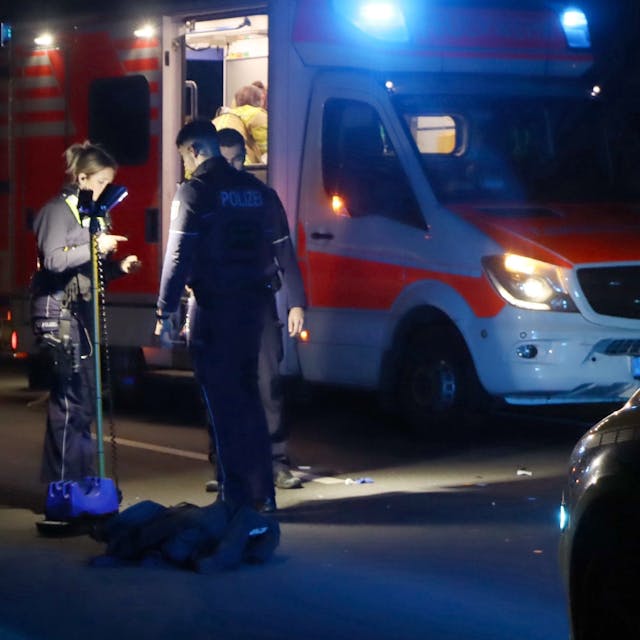Auf dem Foto ist die hell erleuchtete Unfallstelle auf der Rodenkirchener Straße in Wesseling zu sehen. Polizisten sichern Spuren. Im Hintergrund ist ein Rettungswagen zu sehen.