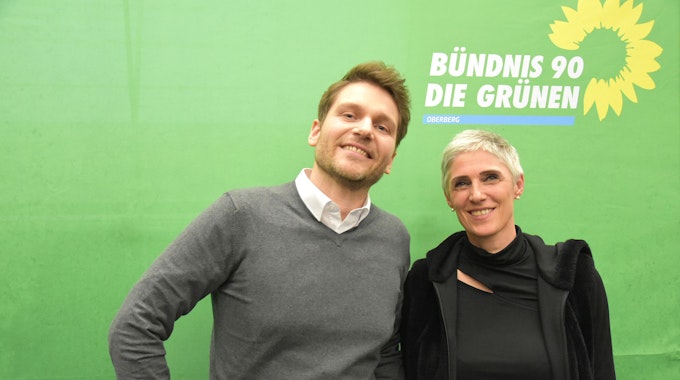 Julian Münster (li.) u. Bernadette Reinery-Hausmann stehen vor einer grünen Wand mit dem Logo der Grünen.