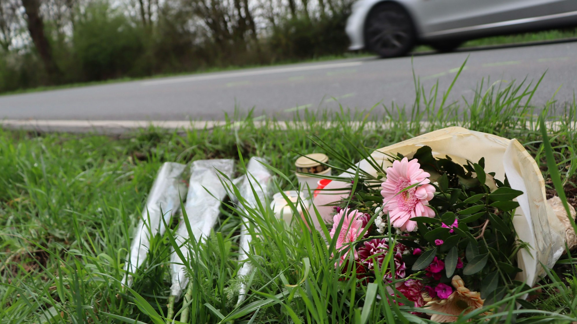 Auf einem Rasensteifen neben einer asphaltierten Staße liegen Blumen, daneben steht ein Grablicht.