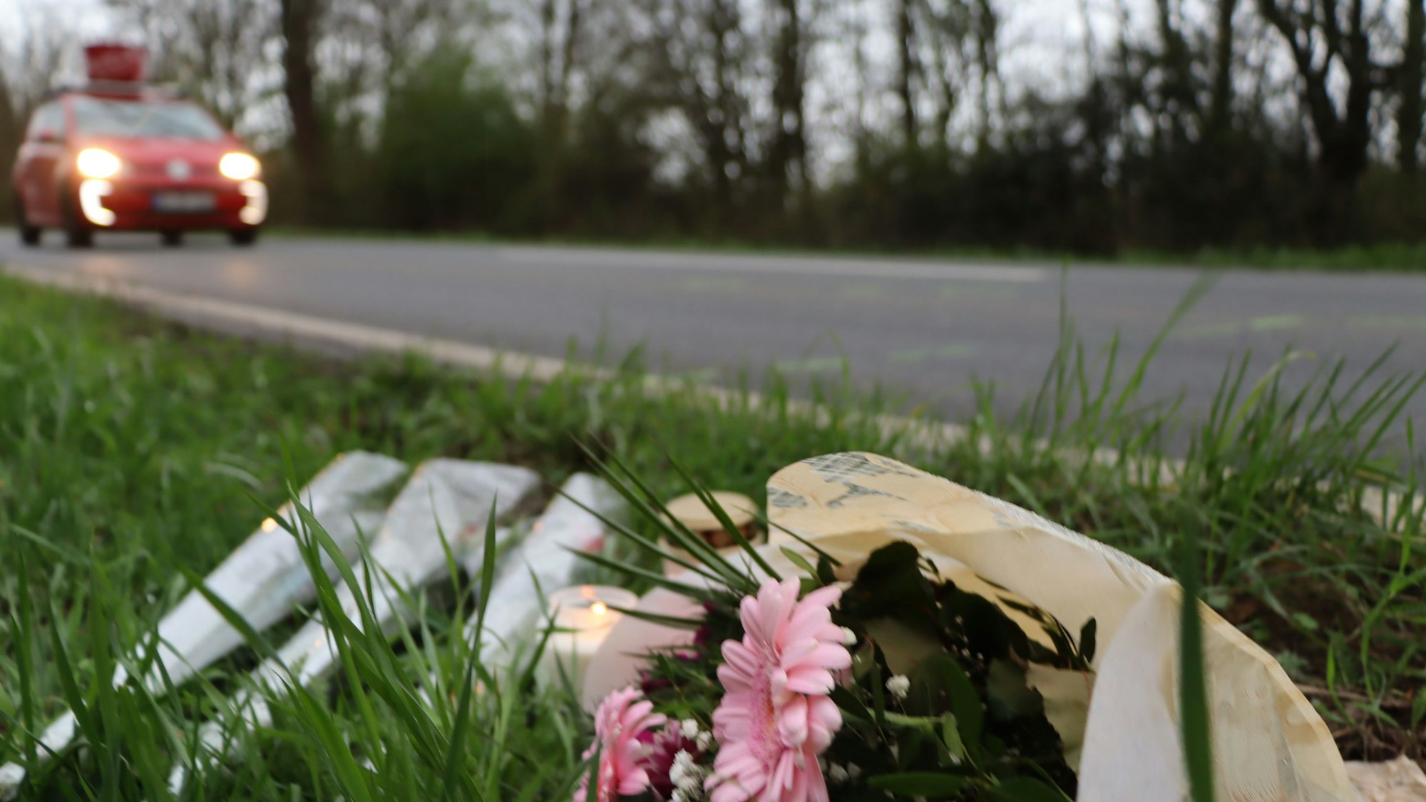 Das Foto zeigt die Unfallstelle in Wesseling. Im Vordergrund sind Blumensträuße und Kerzen zu sehen, im Hintergrund fährt ein Auto vorbei.