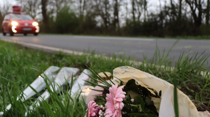 Das Foto zeigt die Unfallstelle in Wesseling. Im Vordergrund sind Blumensträuße und Kerzen zu sehen, im Hintergrund fährt ein Auto vorbei.