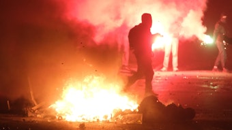 Gegenstände brennen auf einer Kreuzung, zwei vermummte Personen sind zu sehen.