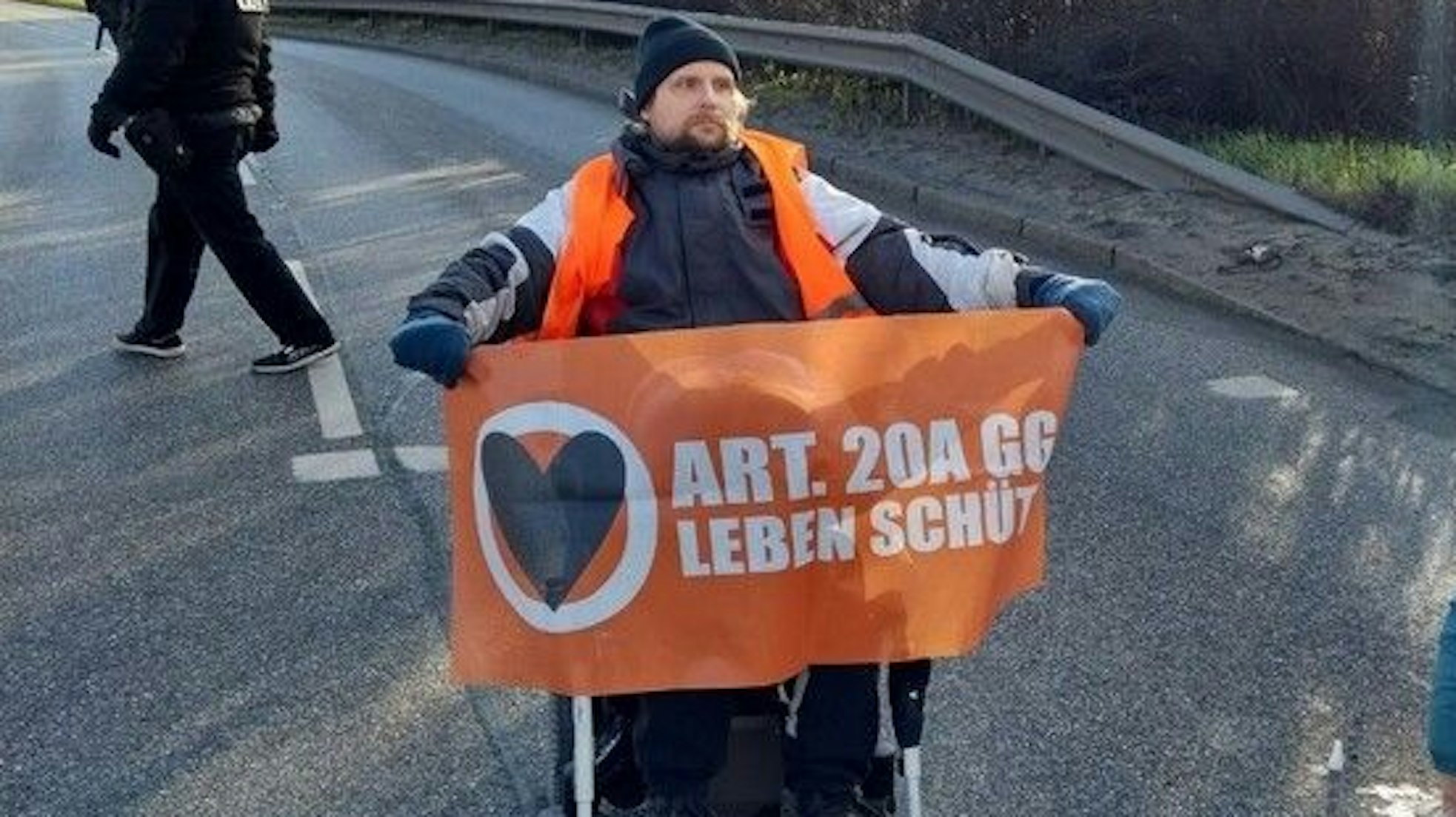Ein Rollstuhlfahrer blockiert am Dienstag (28. März) eine Hauptverkehrsader am Hamburger Hafen. Er hält ein orangefarbenes Banner hoch.