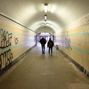 Zu sehen sind zwei Männer, die durch die Unterführung eines Bahnhofs gehen, deren Wände beschmiert sind.