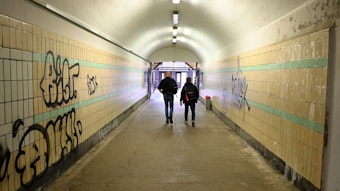 Zu sehen sind zwei Männer, die durch die Unterführung eines Bahnhofs gehen, deren Wände beschmiert sind.