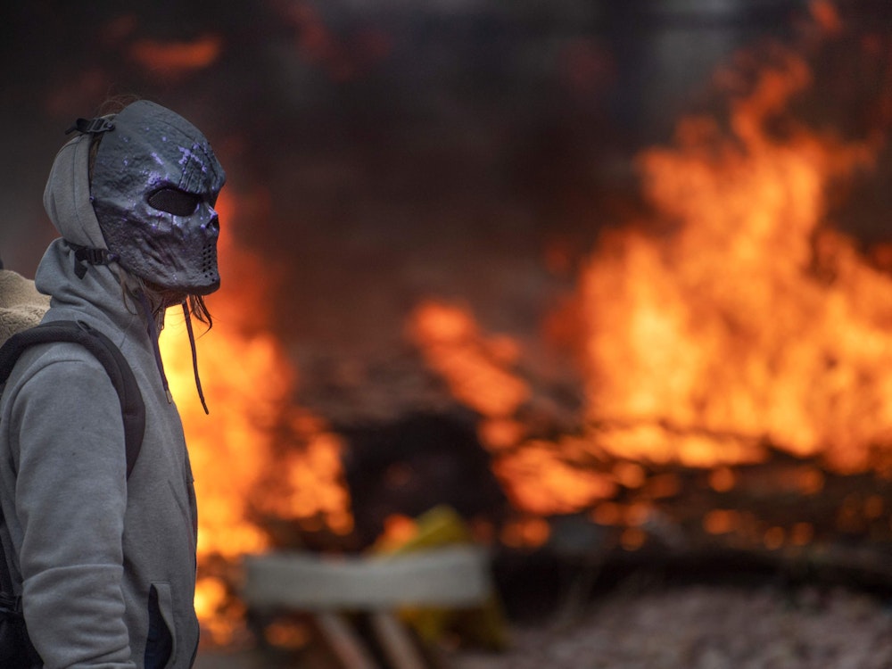 Ein Demonstrant mit Maske steht vor einer brennenden Barrikade, die auf Bahnhofs-Gleisen liegt.