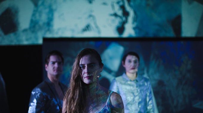 Ein Schauspieler und zwei Schauspielerinnen stehen in blau glänzenden Kostümen auf der Bühne.