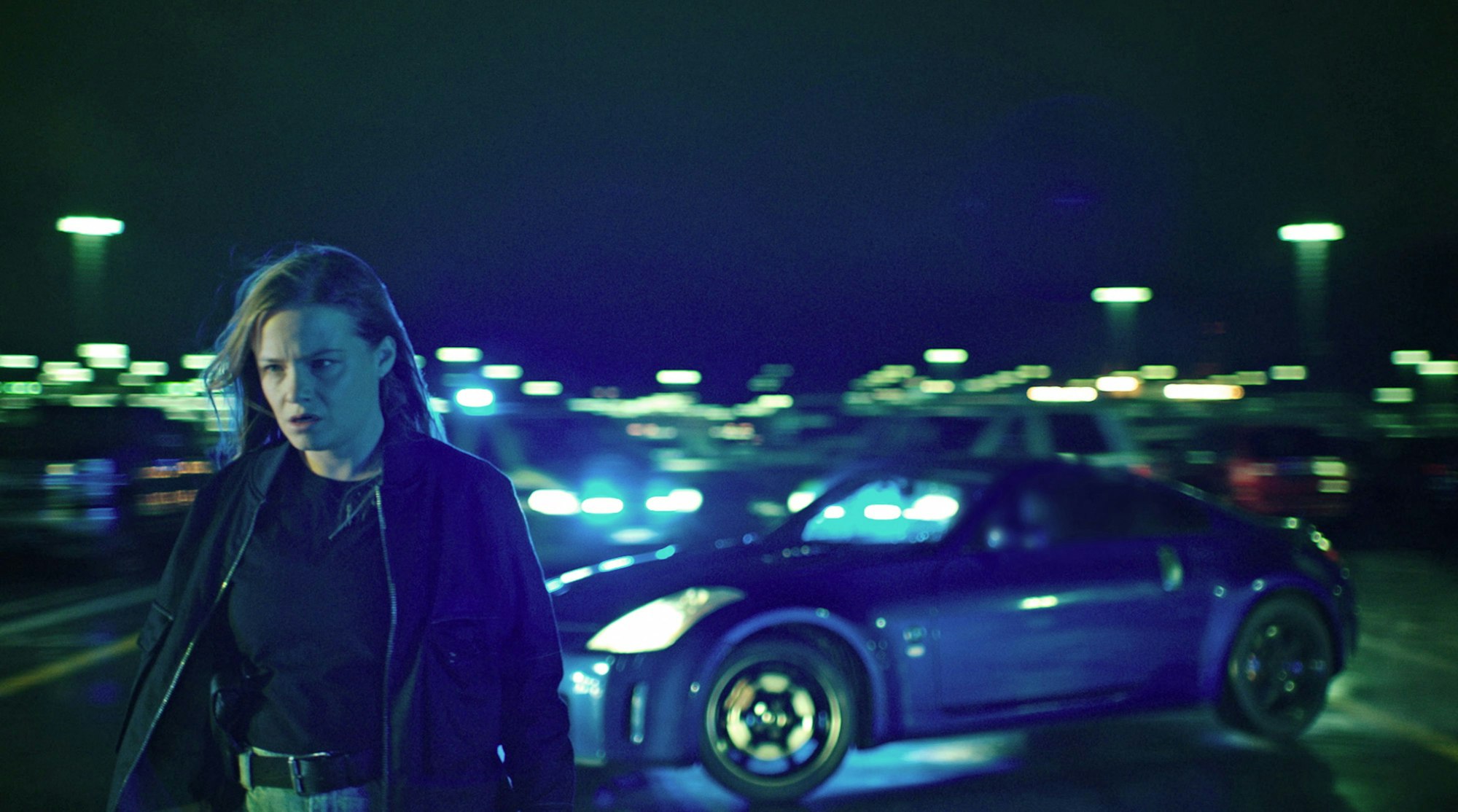 Jasna Fritzi Bauer in der Rolle Liv Moormann im Bremer „Tatort“ „Donuts“. Sie geht nachts von einem Auto weg und schaut düster. Hinter ihr leuchten diverse Lampen sowie die Scheinwerfer des blauen Sportwagens.