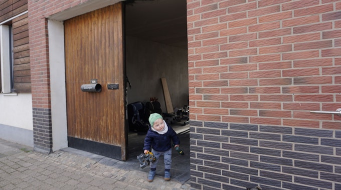 Der kleine Max, einen Spielzeugbagger in der Hand, lugt vorsichtig aus dem Hoftor auf die Straße vor dem Haus.