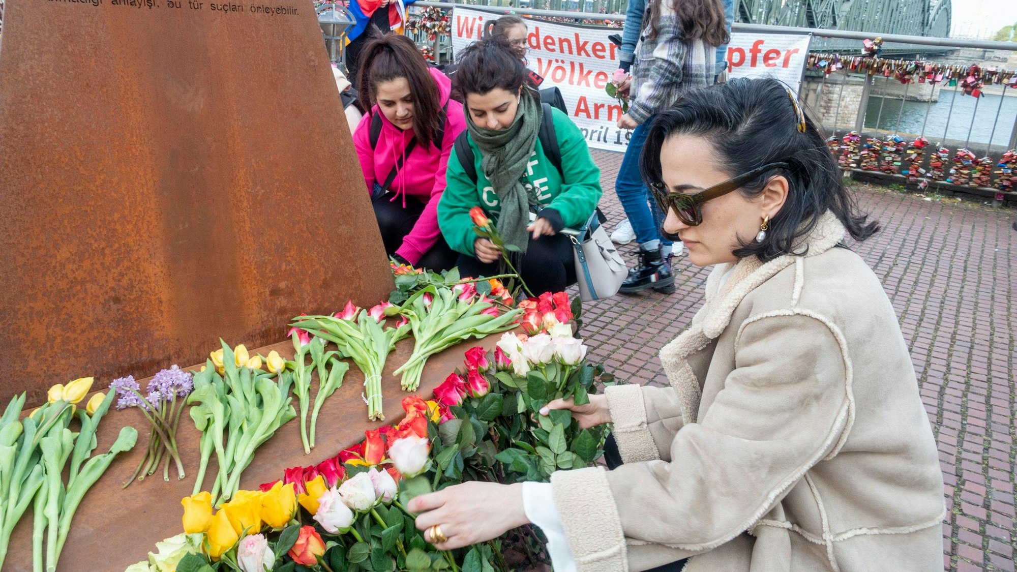 Erinnerung an den Genozid an Armeniern am Heinrich-Böll-Platz: Eine Frau mit Sonnenbrille legt Blumen vor einer Gedenkstele ab. Im Hintergrund ist die Hohenzollernbrücke zu sehen.