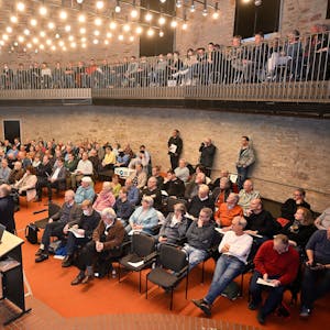 Menschen sitzen im Bensberger Ratssaal und hören einem Sprecher auf der Bühne zu.