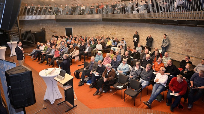 Menschen sitzen im Bensberger Ratssaal und hören einem Sprecher auf der Bühne zu.