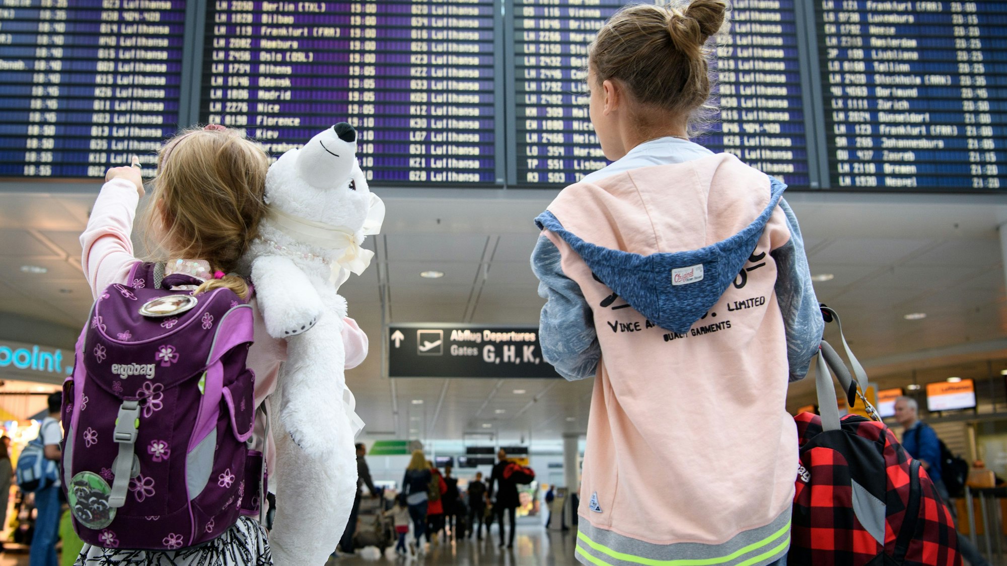Zwei Mädchen blicken auf eine Abflugtafel am Flughafen. Eines trägt einen weißen Teddy im Arm.