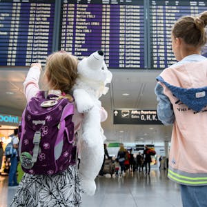 Zwei Mädchen blicken auf eine Abflugtafel am Flughafen. Eines trägt einen weißen Teddy im Arm.