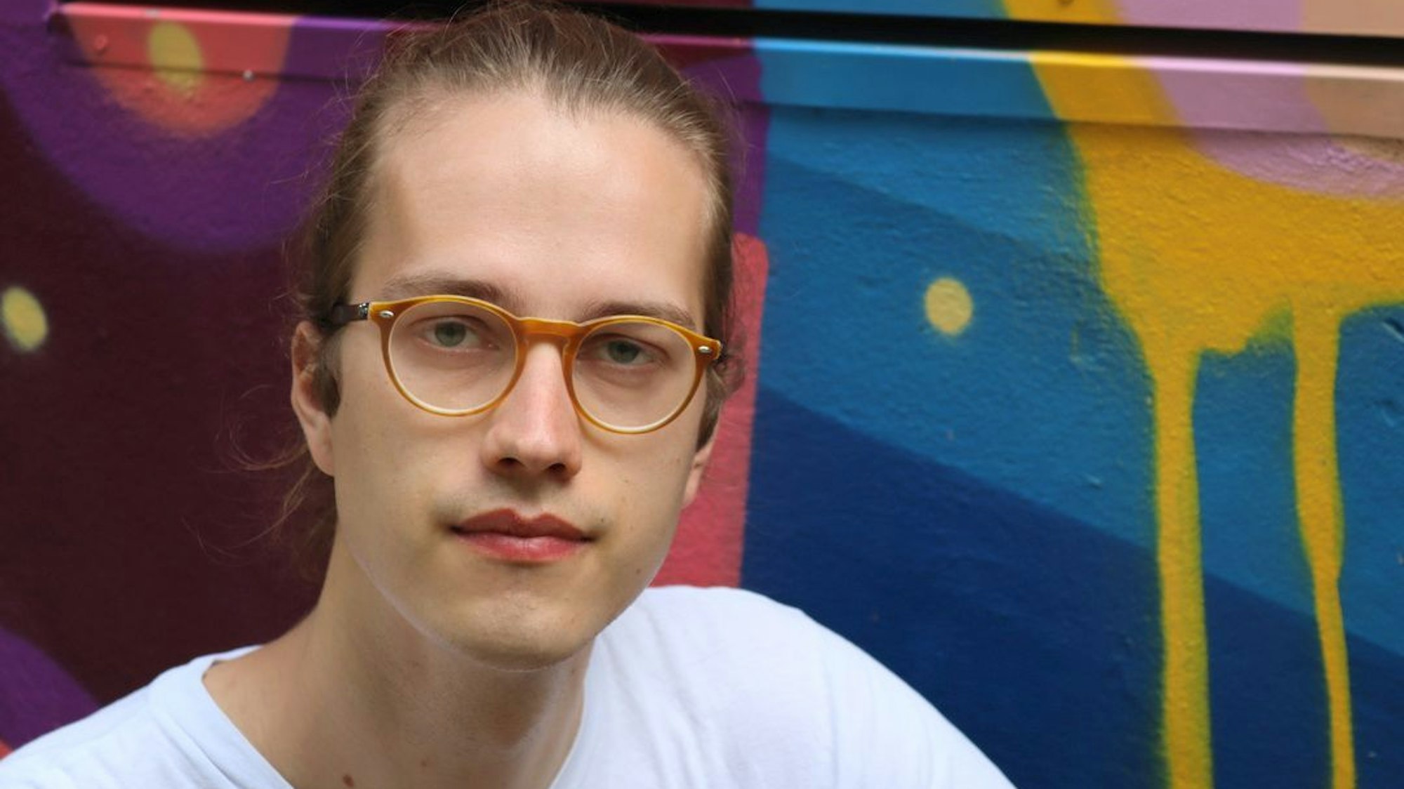 Das Bild zeigt Simon Bahr, er schaut in die Kamera und trägt eine Brille und ein weißes T-Shirt. Im Hintergrund bunte, geometrische Formen an einer Wand.