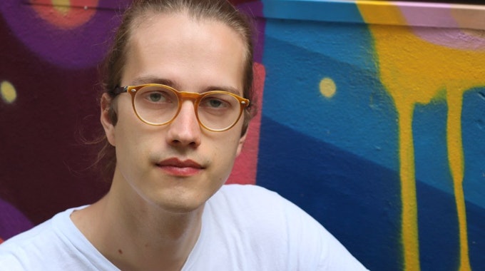 Das Bild zeigt Simon Bahr, er schaut in die Kamera und trägt eine Brille und ein weißes T-Shirt. Im Hintergrund bunte, geometrische Formen an einer Wand.&nbsp;