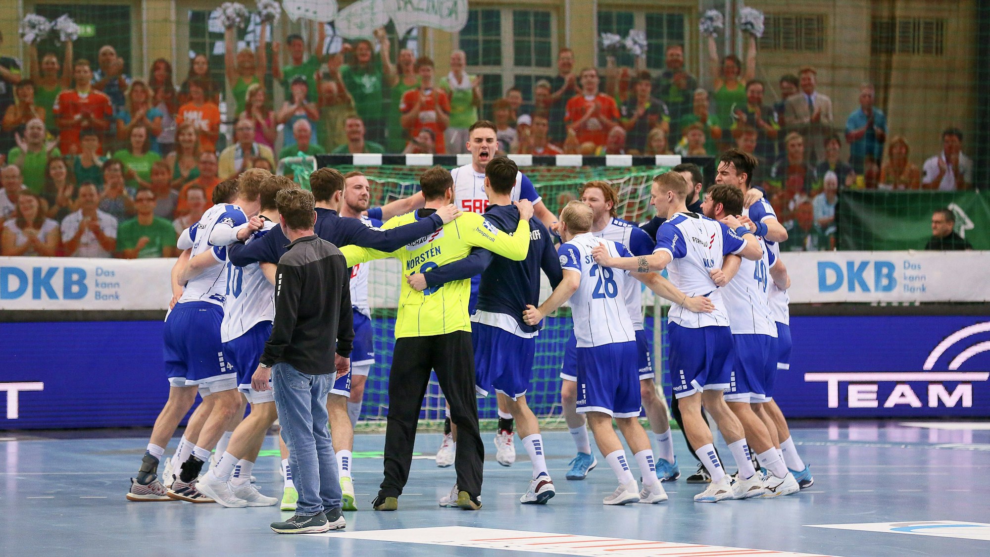 Eine Handballmannschaft steht im Kreis zusammen, die Spieler liegen sich in den Armen und feiert hüpfend den Sieg.