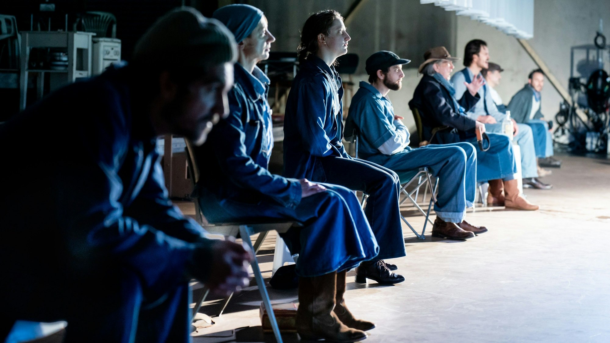 Acht Schauspieler des Kölner Ensembles sitzen, von der Seite fotografiert, nebeneinander auf Klappstühlen im Stück „Früchte des Zorns“ im Depot 2. Sie tragen alle Kostüme aus Jeansstoff.