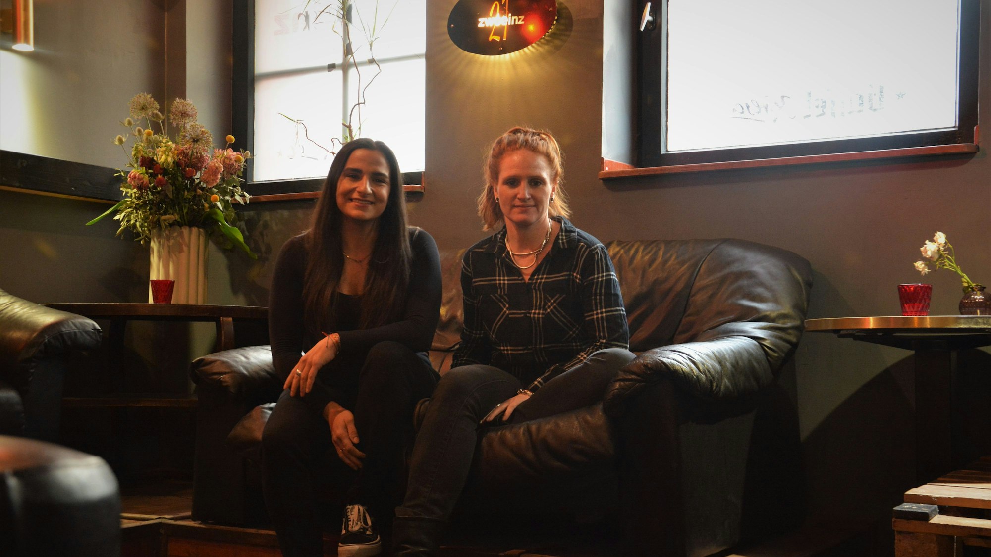 Walentini Malliou und Jana Voss sitzen auf einer Couch in der Bar ZwoEinz, über ihnen ist das Logo der Bar zu sehen.